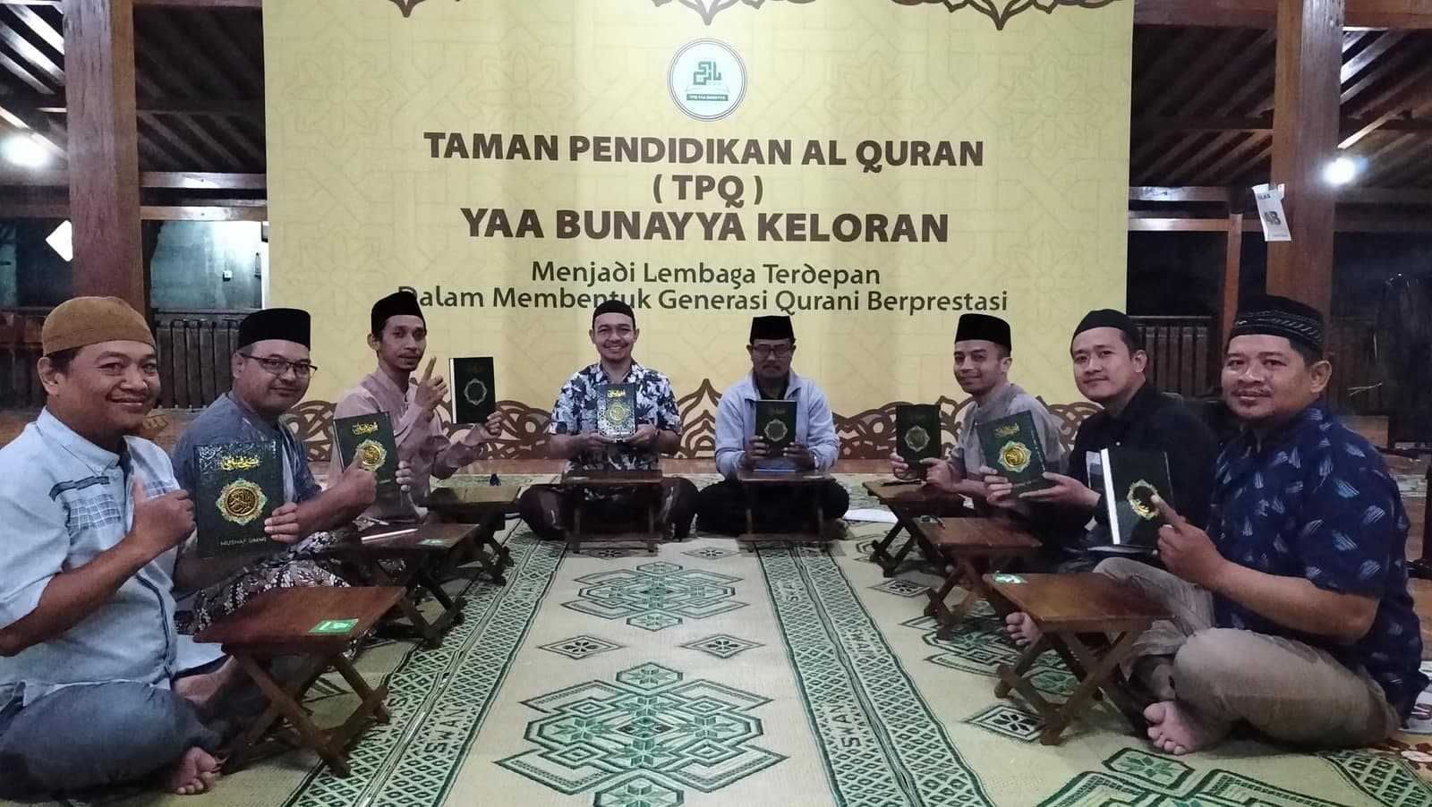 Para ustadz memegang buku pembelajaran di depan banner TPQ